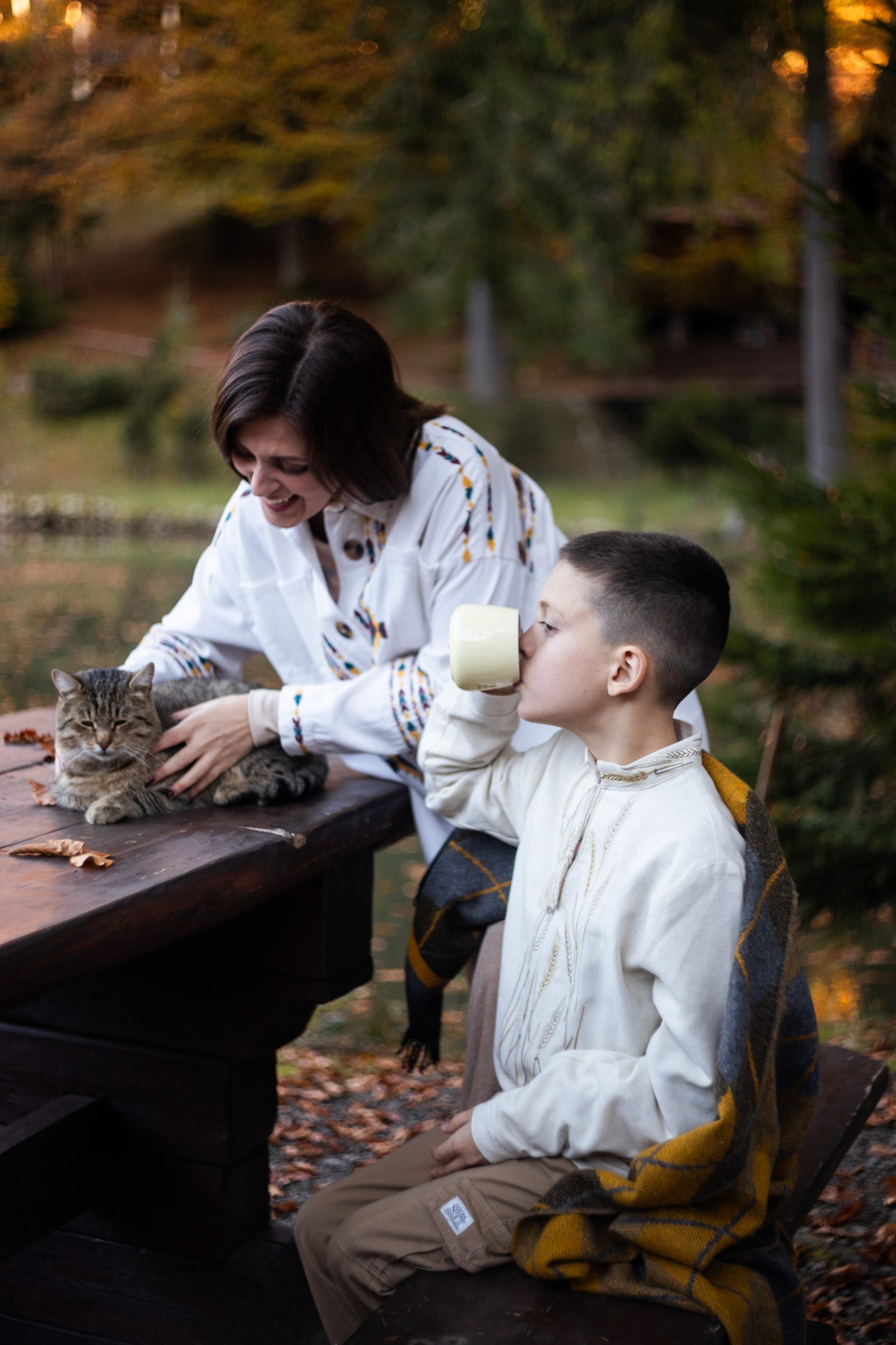 Mama z synem piją herbatę pod kocem przy stole wśród jesiennego lasu, obok kot